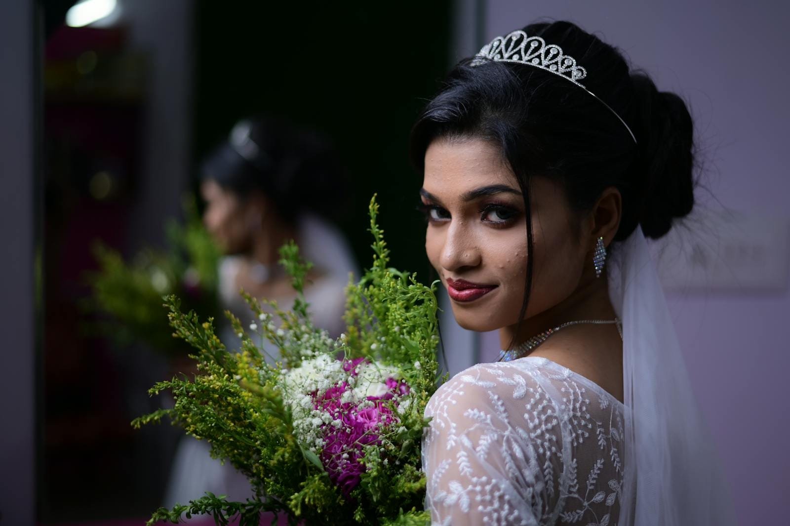 A stunning bride with a bouquet poses elegantly indoors, showcasing her captivating wedding attire and tiara.