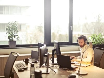 A businessman sits at a desk using multiple computers and a headset in a well-lit modern office.