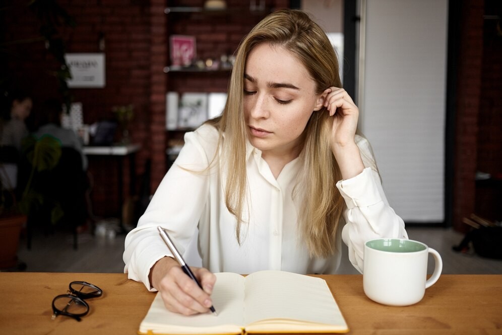 candid-shot-attractive-blonde-student-girl-white-blouse-doing-homework-workplace-home-writing-down-open-copybook-drinking-tea-having-serious-concentrated-facial-expression_343059-4187