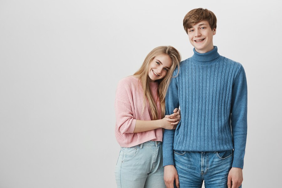 cheerful girl with long blonde hair pink sweater with braces laughing hugging fair haired boyfriend Straipsniai.lt