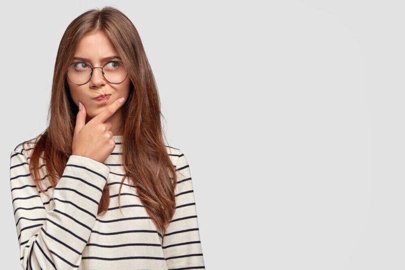 confused-lovely-female-teenager-holds-chin-looks-thoughtfully-aside-has-dark-hair-wears-striped-sweater-isolated-white-wall_273609-16546