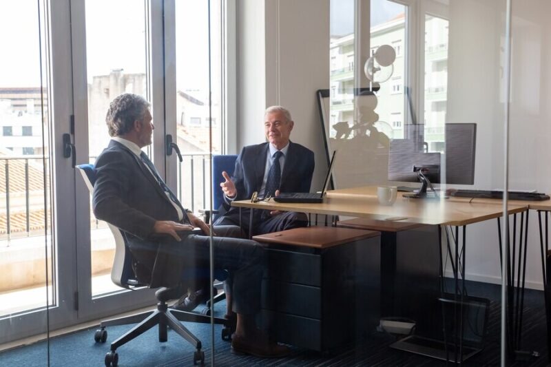 friendly-mature-business-partners-meeting-office-sitting-workplace-with-laptop-talking-view-through-glass-wall-business-portrait-concept_74855-14245