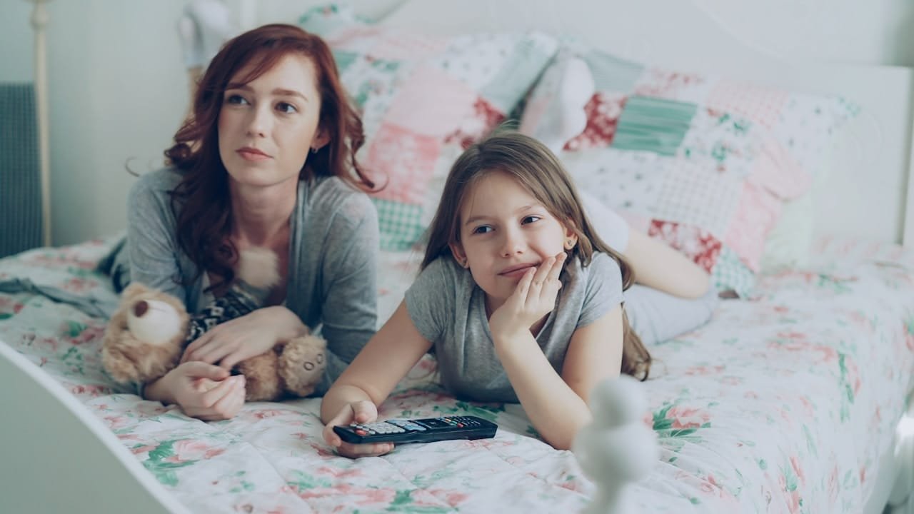 Mother and daughter watch television together on the bed.