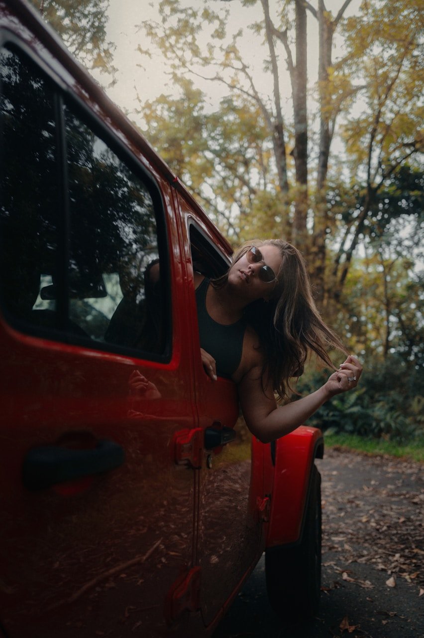 Woman poses from a red jeep in the woods.