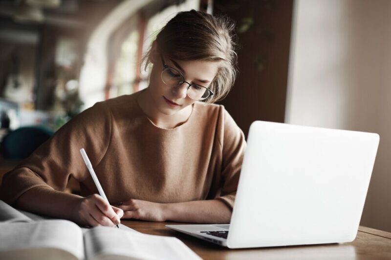 hardworking-focused-woman-trendy-glasses-concentrating-writing-essay-sitting-cozy-cafe-near-laptop-working-making-notes-carefully_197531-21320