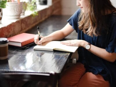 woman sitting in front of black table writing on white book near window