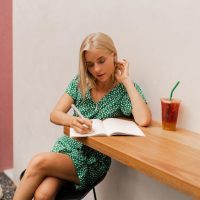 pretty-blond-woman-writing-notebook-wearing-summer-dress-sitting-modern-cafe-with-white-walls_273443-5258