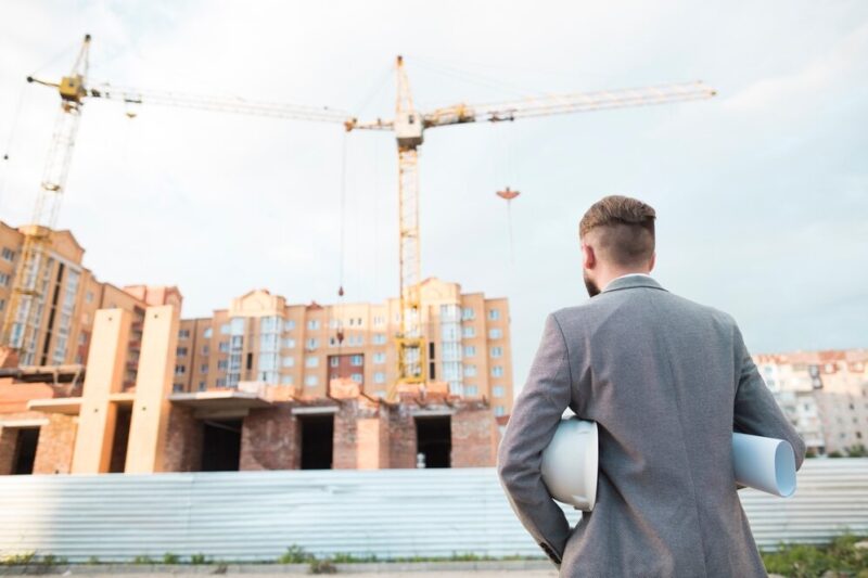 rear-view-male-architect-holding-blueprint-hard-hat-looking-construction-site_23-2148203954