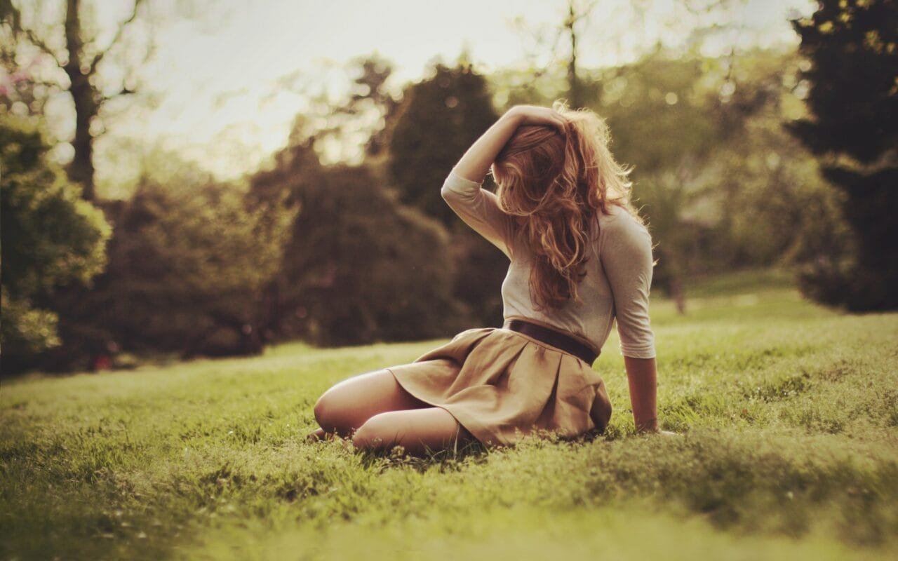Sad Girl Sitting In Meadow With Long Curly Hair Touching Of Her Head With Her Right Hand