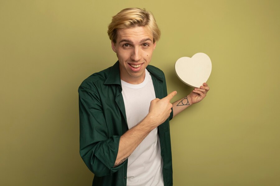 smiling young blonde guy wearing green t shirt holding points heart shape Straipsniai.lt