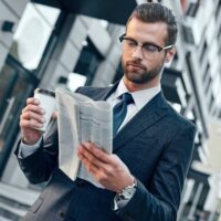 young-businessman-suit-glasses-holding-paper-cup-reading-business-newspaper