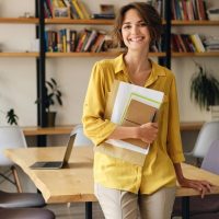 young-cheerful-woman-yellow-shirt-leaning-desk-with-notepad-papers-hand-while-joyfully-looking-camera-modern-office_574295-3824