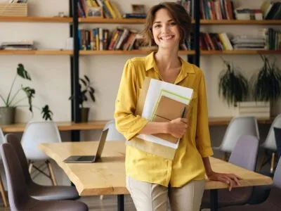young-cheerful-woman-yellow-shirt-leaning-desk-with-notepad-papers-hand-while-joyfully-looking-camera-modern-office_574295-3824