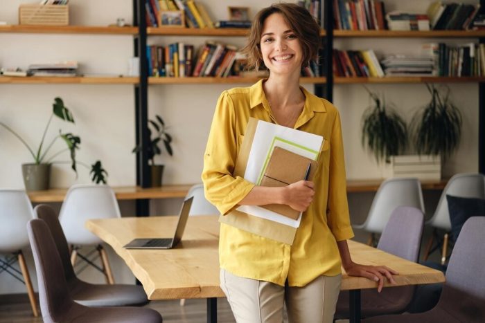 young-cheerful-woman-yellow-shirt-leaning-desk-with-notepad-papers-hand-while-joyfully-looking-camera-modern-office_574295-3824