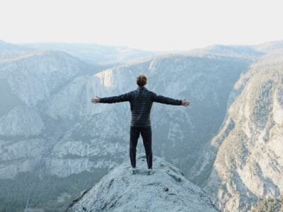 man opening his arms wide open on snow covered cliff with view of mountains during daytime